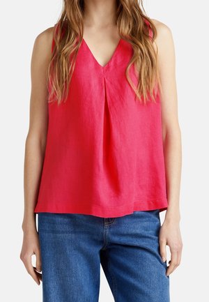 Woman wearing a sleeveless red V-neck top with front pleat paired with blue jeans, shown from neck to hips against a plain background.