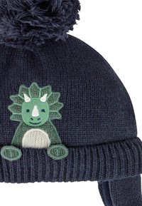 Căciulă tricotată de culoare bleumarin, cu o aplicație verde în formă de dinozaur în față, decorată cu un pompon pufoșel. Textură moale, cu urechi pentru căldură.