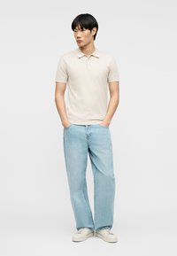 Homme portant un polo à manches courtes beige clair, un jean larges bleu clair et des baskets blanches, debout les mains dans les poches.