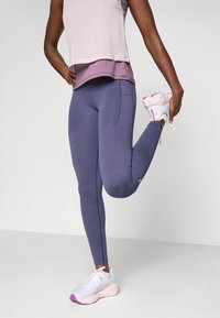Purple athletic leggings with a textured design and side pocket, paired with a light purple layered tank top and white-pink running shoes.