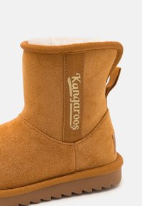 Bottines en daim marron avec une douce doublure crème, une patte arrière et une semelle en caoutchouc texturée. Présente le logo doré "Kangaroos" sur le côté.