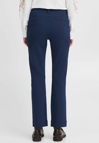 Marineblaue Hose mit geradem Bein, Gesäßtaschen, dezenten Falten und einer glänzenden Textur, kombiniert mit dunklen Schuhen.