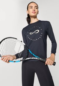 Langermet svart treningsskjorte med et sentrert grafisk design og rund hals, kombinert med svarte tights. Holder et tennisrackett med blå detaljer.