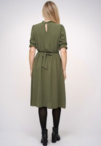 Rochie midi verde măsline cu mâneci scurte puff, talie strânsă și detaliu de decolteu în formă de cheie pe spate. Purtați-o cu cizme negre până la gleznă și colanți.
