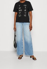 Camiseta de algodón negra con diseño de texto blanco, combinada con jeans de pierna ancha de color azul claro. Lleva sandalias de tacón negras y un bolso negro.