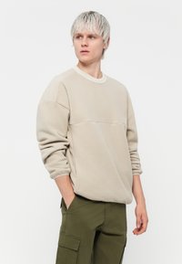 Sweatshirt en polaire beige avec un col rond, des poignets côtelés et un détail de couture horizontal, associé à un pantalon cargo vert olive.