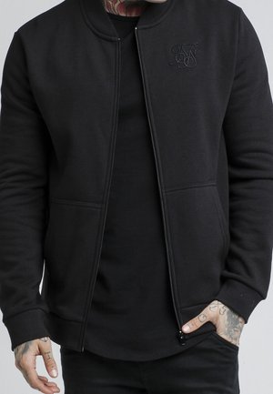 Homme portant une veste noire zippée sur une chemise noire avec des mains tatouées, une main dans la poche, fond clair uni.