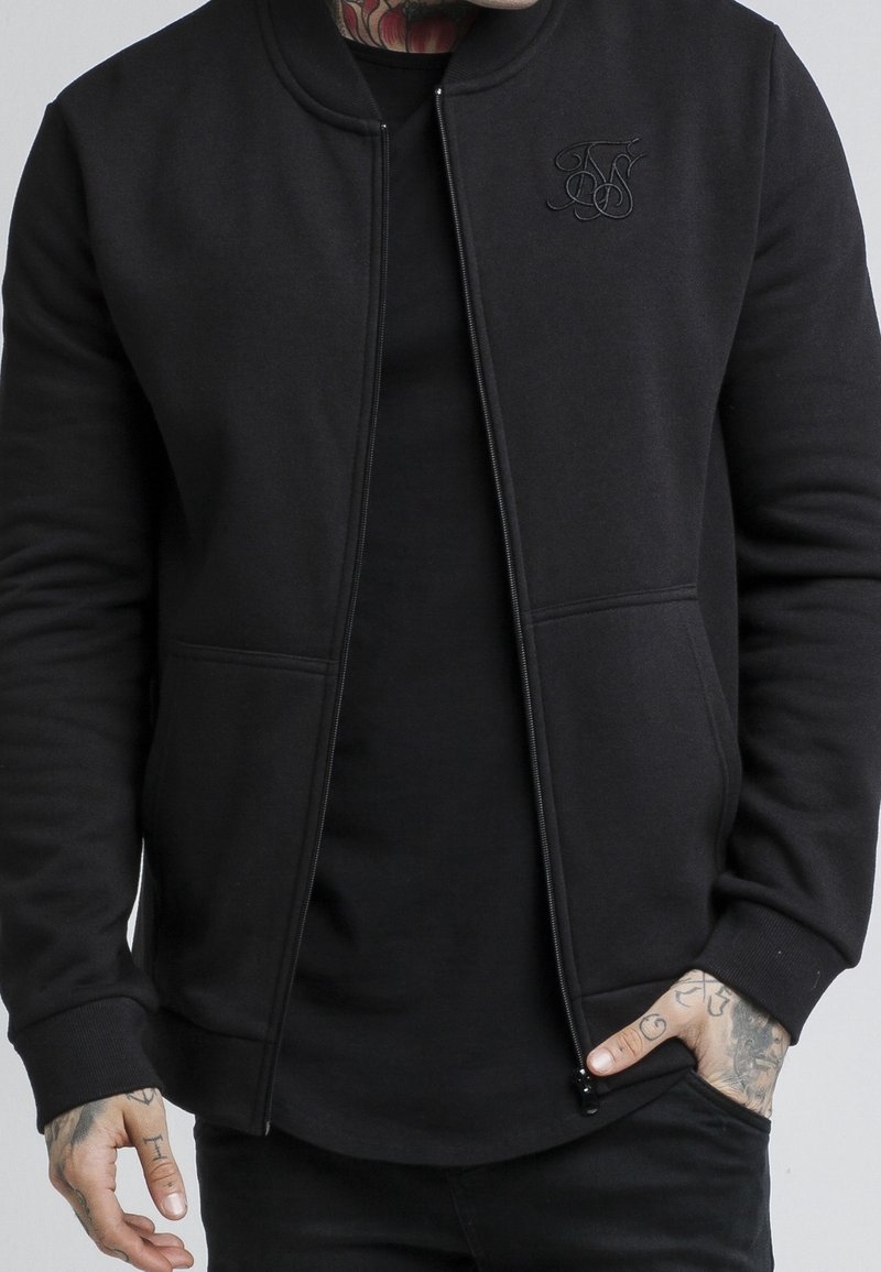 Homme portant une veste noire zippée sur une chemise noire avec des mains tatouées, une main dans la poche, fond clair uni.