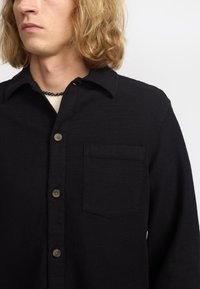 Camisa negra de botones hecha de tela texturizada, con un bolsillo en el pecho, cuello puntiagudo y botones marrones. Diseño sencillo con un corte relajado.