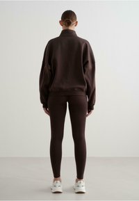 Sudadera marrón de gran tamaño con cuello alto, combinada con leggings marrones a juego. El modelo lleva zapatillas blancas. Textura suave, diseño minimalista.