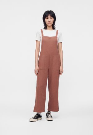 Persona in piedi di fronte, indossa una t-shirt bianca, una salopette marrone larga e sneakers nere su uno sfondo bianco semplice.