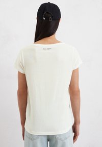 Lichte crèmekleurige t-shirt met korte mouwen en ronde hals, met een subtiele logo-afdruk op de achterkant en een relaxed fit, gecombineerd met blauwe jeans.