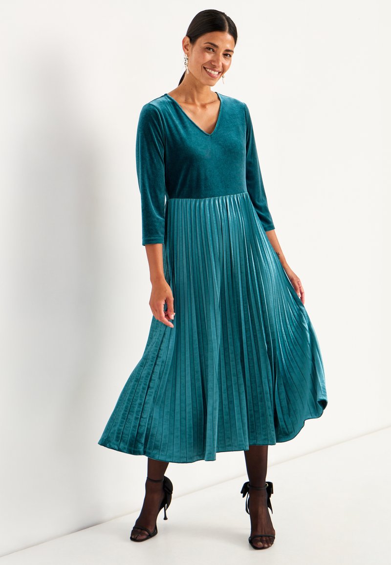 Teal fluwelen jurk met een V-hals, driekwartmouwen en een geplooide rok. Gladde textuur met een vloeiende ontwerp en een lengte tot de enkels.