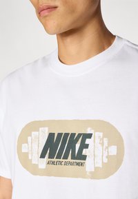 Biała bawełniana koszulka z dużą szaro-beżową grafiką z napisem "NIKE" i "DZIAŁ SPORTOWY" oraz ikoną podnoszenia ciężarów.