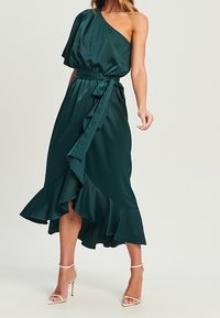 Vestido de un solo hombro, de color verde oscuro, hecho de un tejido suave. Presenta un cinturón de lazo, un dobladillo con volantes y un diseño asimétrico.