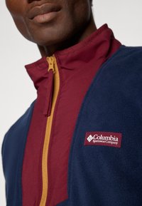 Giacca in pile di colore blu navy e bordeaux con zip dorata. Presenta il logo della Columbia Sportswear Company sul lato sinistro. Texture morbida.