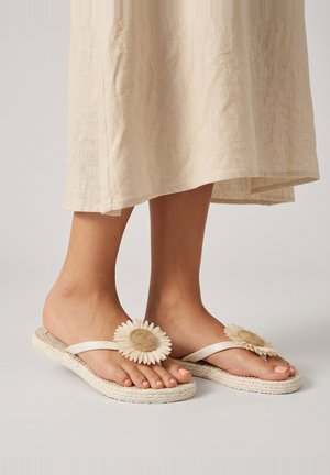 Feet wearing beige sandals with large fabric sunflower decorations and braided soles, standing on a plain surface with a light skirt.