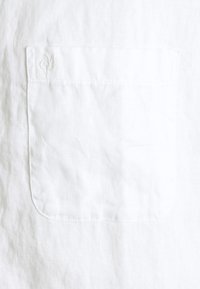 Camicia bianca in lino caratterizzata da una tasca sul petto con una leggera texture e dettagli cuciti, completata da un logo impresso nell'angolo.