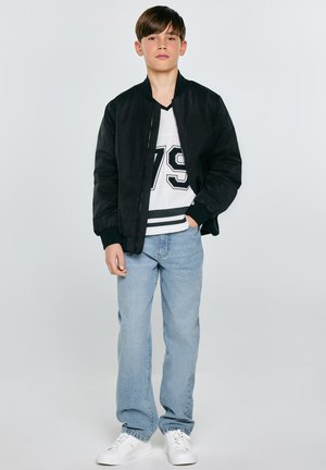 Chlapec stojí, má na sobě černou bundu, bílou košili s číslem 79, světle modré džíny a bílé tenisky, ruka v kapse.