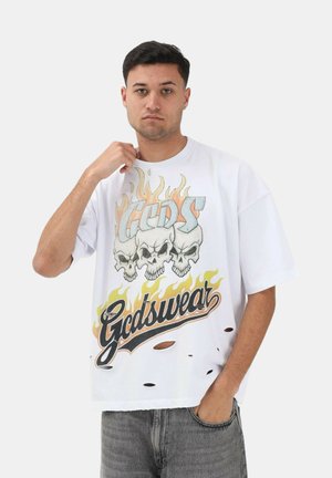 Giovane uomo che indossa una maglietta bianca oversize con teschi infuocati e testo "Gcdswear", mano vicino al colletto, in piedi contro uno sfondo semplice.