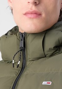 Olivgrüne Steppjacke mit hohem Kragen, Frontreißverschluss und verstellbarem Kordelzug. Verfügt über ein kleines Logo auf der Brust.