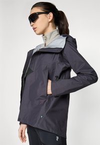 Donkergrijze jacket met een lichtgewicht ontwerp, voorzien van een capuchon, ritssluitingen en een contrasterende lichtgrijze voering. Over het geheel een gladde textuur.