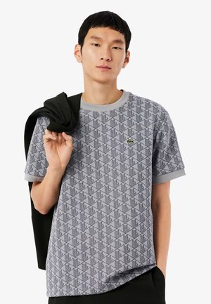 Jeune homme portant un t-shirt gris à motifs Lacoste, tenant une veste noire sur son épaule devant un fond uni.