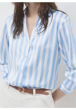 Femme portant une chemise à rayures verticales bleu clair et blanc, boutonnée et rentrée dans un pantalon blanc avec une ceinture tressée marron.