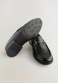 Sorte læderloafers med en glat overflade, der har dekoreret syning og en sølvfarvet hardwaredetalje. Gummisål med cirkulært grebs mønster.