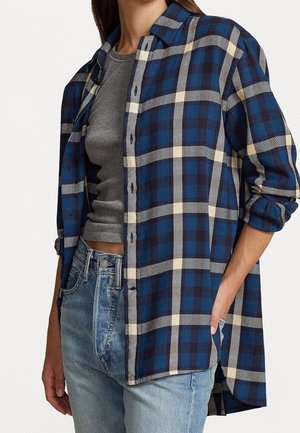 Femme portant une chemise à carreaux bleue et beige boutonnée sur un crop top gris et un jean bleu clair, avec la main droite dans sa poche.