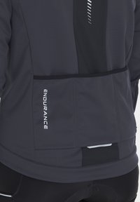 Grå sportjacka med en svart panel, reflektiva detaljer och en ficka med dragkedja. Tygtexturen verkar slät och hållbar.