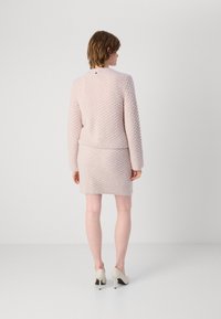 Lichtroze, gebreide top met textuur, lange mouwen en een aansluitende bijpassende rok. Geaccentueerd met een klein logo op de achterkant. Klassieke hakken maken de look compleet.