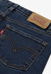 Vaqueros de mezclilla de color azul oscuro, con costuras visibles, un parche de marca de cuero y un diseño distintivo en el bolsillo trasero.
