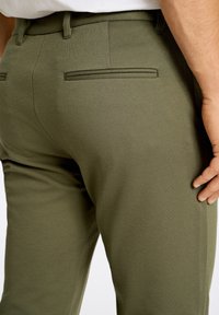 Pantaloni tailored verde oliva con una texture liscia, dotati di due tasche posteriori con dettagli cuciti e una vita a media altezza.