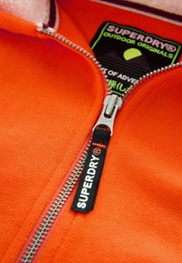 Oranžová bunda na zips s kovovým zipsom, obsahujúca čiernu nálepku s nápisom "SUPERDRY" a farebnými značkovými detailmi. Viditeľná mäkká textúra.