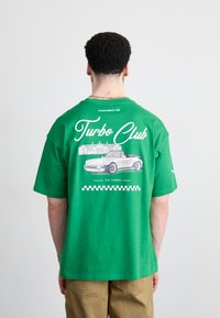 Grøn bomulds t-shirt med en hvid grafik af en cabriolet og teksten "Turbo Club" samt et ternet mønster i bunden.