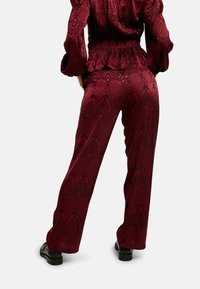 Bordové vzorované nohavice s širokými nohavicami a textúrovaným povrchom. Pás je nariasený a nohavice sú skombinované s čiernou obuvou.