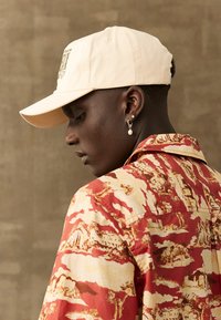 Casquette de baseball beige et chemise rouge à motif présentant une impression de paysage, avec une texture de tissu lisse et des tons terreux contrastants.