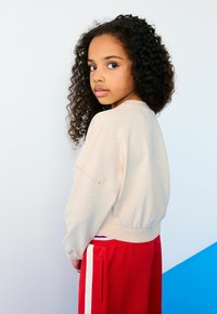 Helles rosa Sweatshirt mit Rundhalsausschnitt und geripptem Saum, kombiniert mit roten Jogginghosen mit weißen Seitenstreifen. Rückansicht mit lockigem Haar.