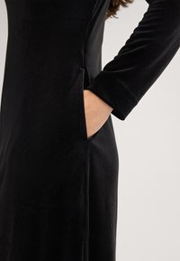Schwarzes Samtkleid mit langen Ärmeln, Seitentasche und einer glatten Textur. Verfügt über eine taillierte Silhouette und einen dezenten Glanz.