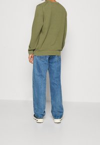 Osoba ubrana w zieloną koszulę z długim rękawem, luźne niebieskie dżinsy i białe trampki, stojąca tyłem do gładkiej białej ściany.