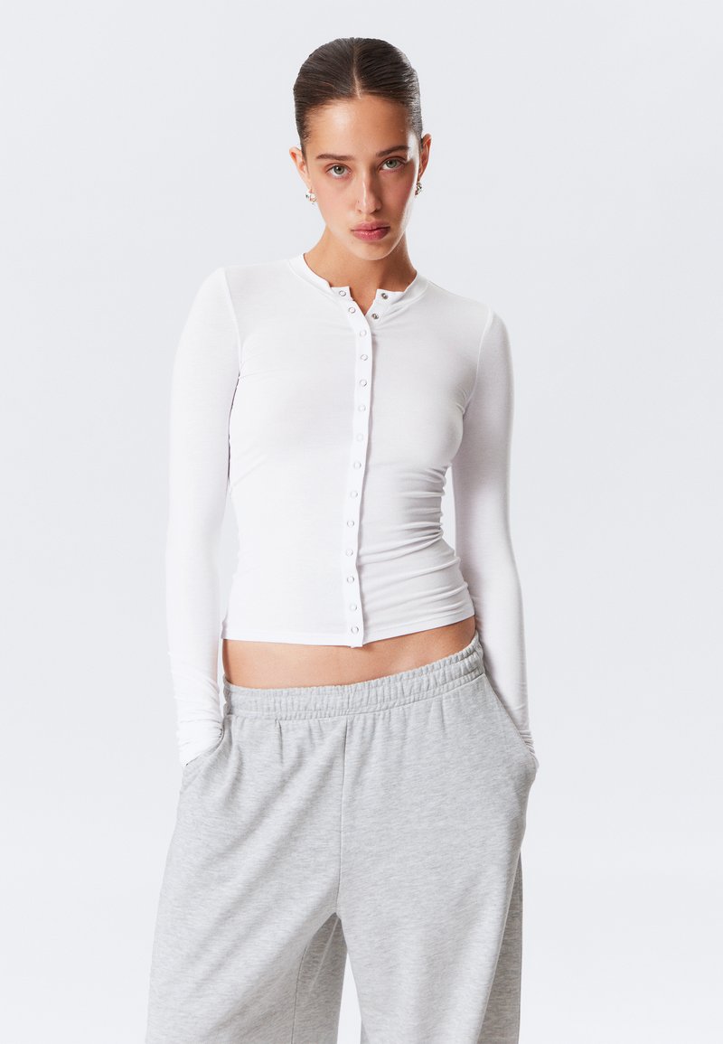 Weißes Langarmshirt mit Rundhalsausschnitt, Knopfleiste und schmaler Passform, kombiniert mit grauen Sweatpants mit elastischem Bund.