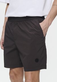 Shorts marron foncé en tissu léger, dotés d'une taille élastique, de poches latérales et d'un patch logo au bas.
