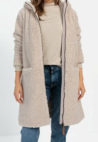 Manteau long en fausse fourrure beige avec capuche, fermeture éclair frontale et deux poches latérales, porté sur un haut gris clair et un jean bleu. Tissu doux et texturé.