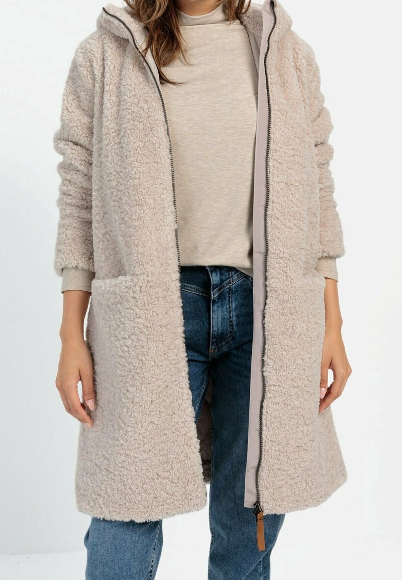 Manteau long en fausse fourrure beige avec capuche, fermeture éclair frontale et deux poches latérales, porté sur un haut gris clair et un jean bleu. Tissu doux et texturé.