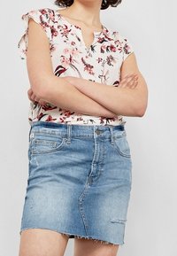 Květinová halenka s krátkými rukávy v bílé barvě s růžovými a červenými vzory, doplněná světle modrou džínovou sukní s třepeným lemem a mírným opotřebením.