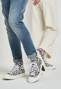 Zapatillas altas con un estampado de leopardo en blanco y negro, cordones blancos, suelas de goma y logotipos de contraste. Jeans de corte recto con dobladillo visible.