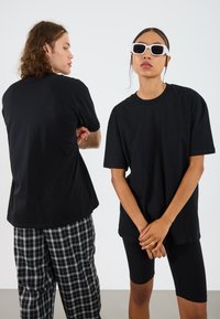 T-shirt noir surdimensionné présenté sur deux modèles ; l'un portant un pantalon à carreaux, l'autre un short de cyclisme noir, avec une texture lisse et simple.