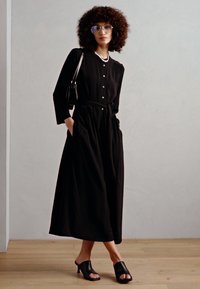 Abito maxi nero con bottoni e maniche lunghe, vita arricciata e tasche; abbinato a mule nere con punta aperta e una piccola borsa.