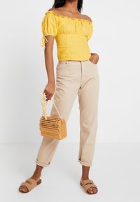 Top giallo off-the-shoulder con maniche a palloncino, jeans beige a vita alta, borsa in legno intrecciato e sandali scivolati marroni.
